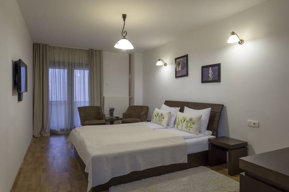 Une chambre à coucher avec un lit, deux chaises, une télévision et une fenêtre avec des rideaux.