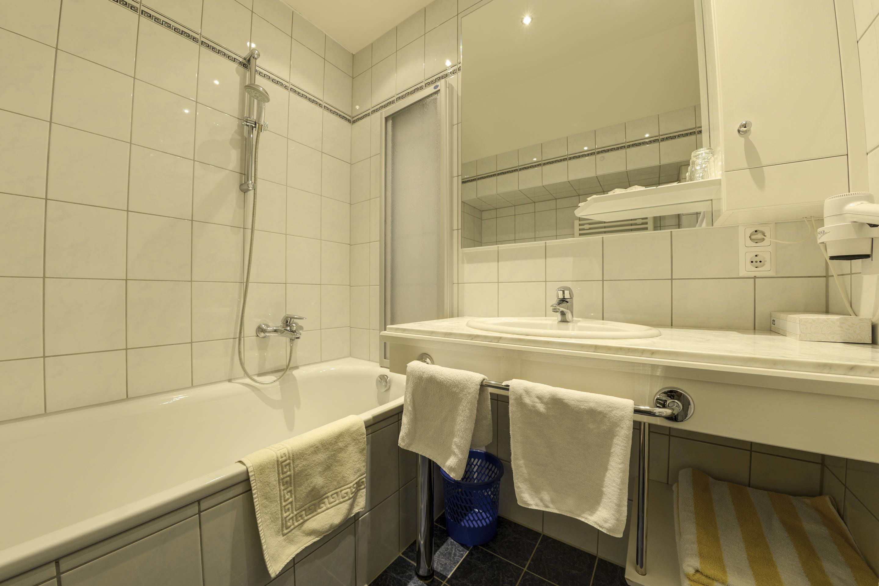 Un baño moderno con bañera, lavamanos y espejo. Hay toallas colgadas en la pared y una cesta azul cerca del lavamanos.