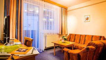 Et hotelværelse med sofa, et lille bord, en stol, et skrivebord med telefon og et vindue med gardiner.