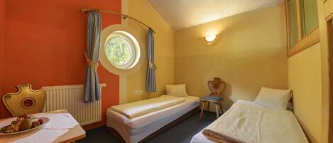 A hotel room with two single beds, a round window, a table with a plate of food, and a chair.