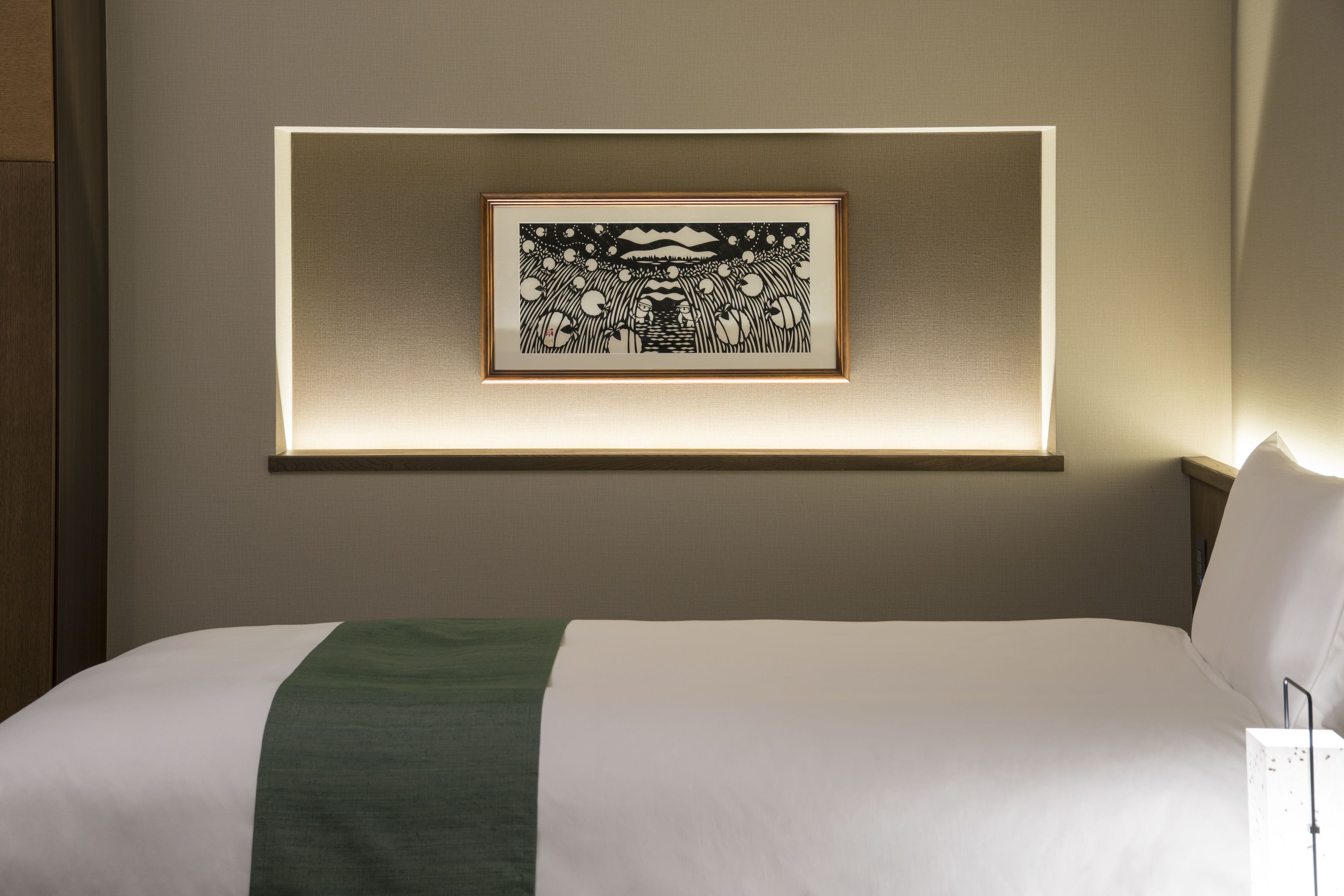 Ein ordentlich bezogenes Bett mit weißgrüner Tagesdecke, ein gerahmtes Kunstwerk darüber und ein Nachttisch mit Lampe.