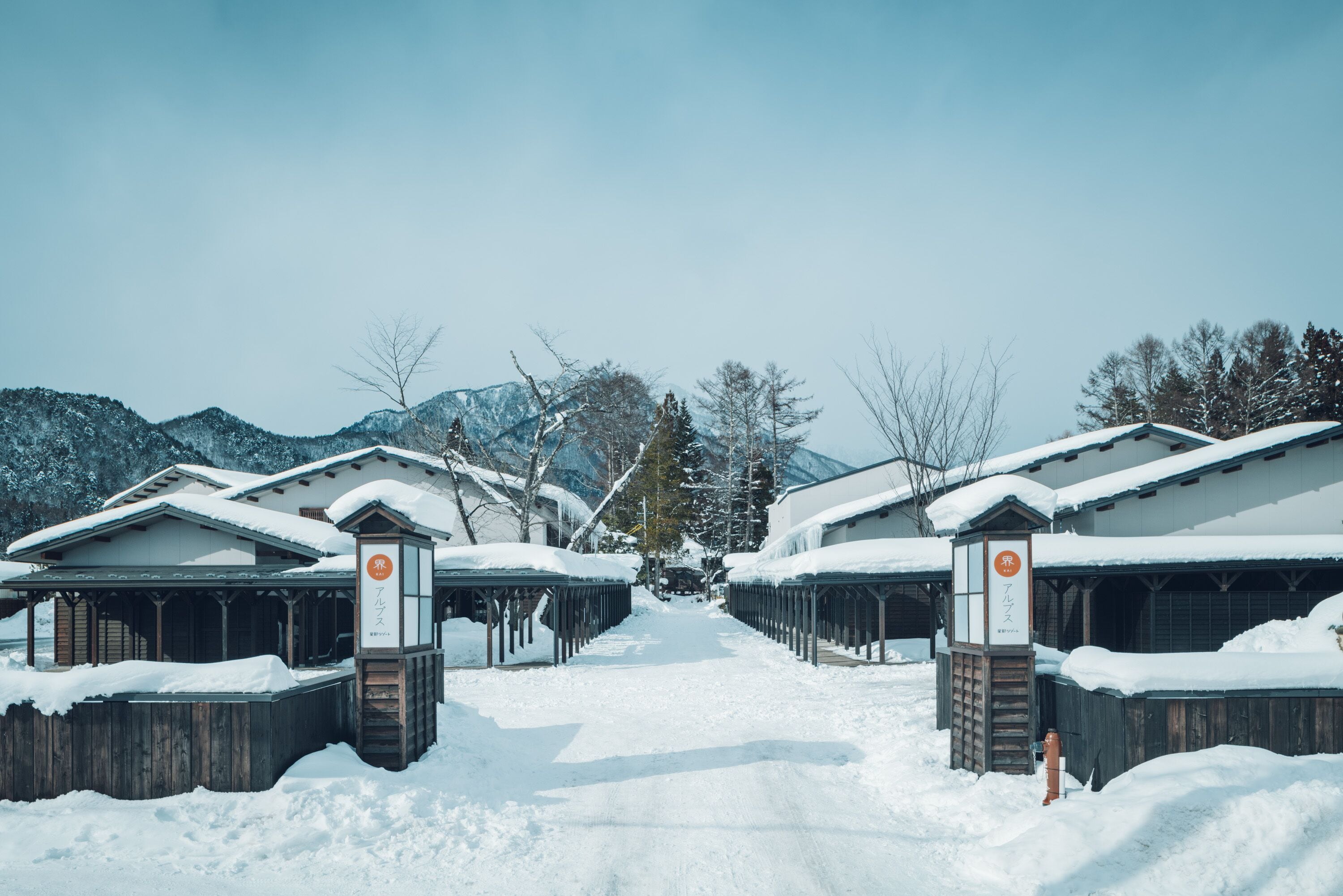 A snow-covered pathway leading to a row of traditional wooden buildings with snow-covered roofs and a clear sky.