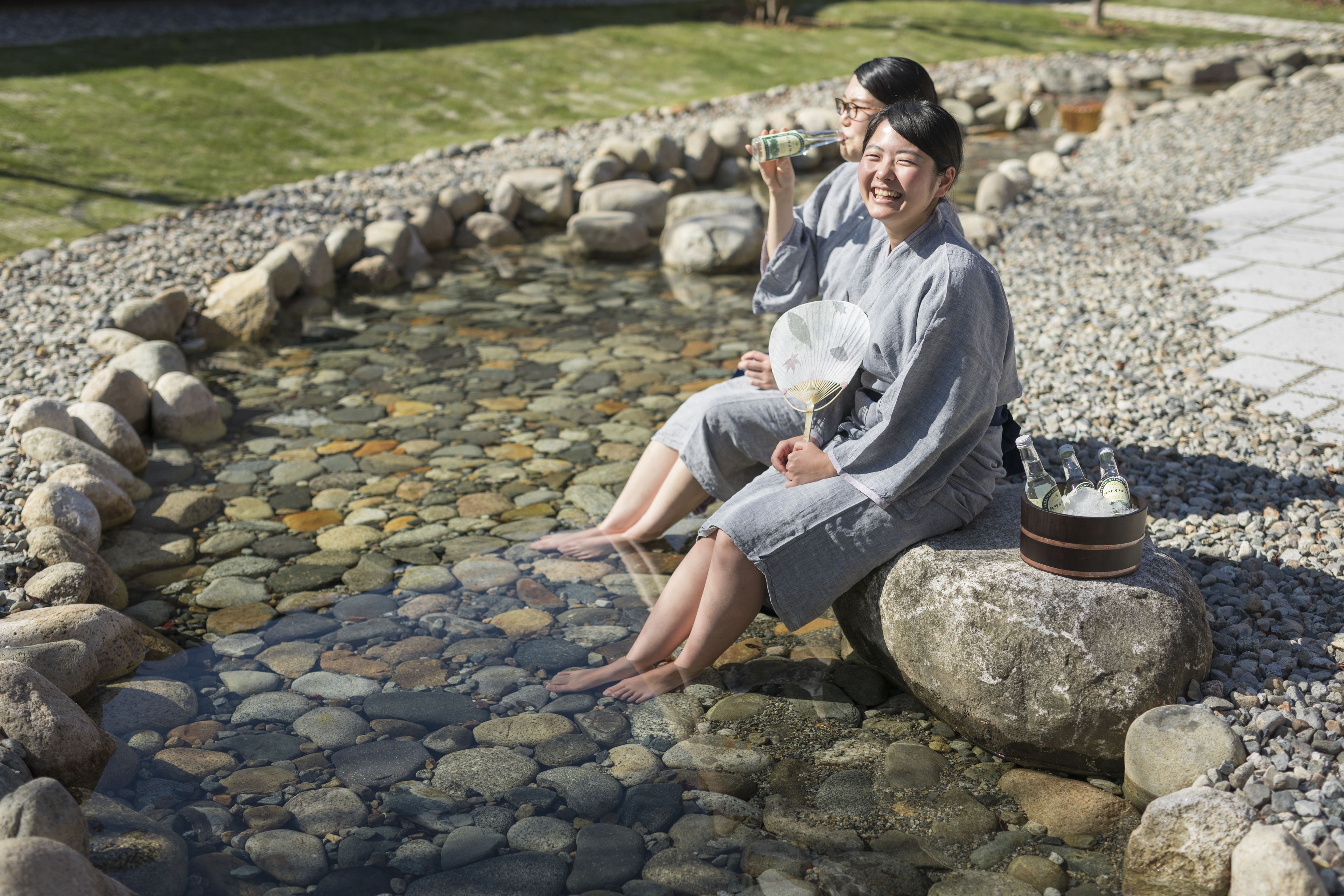 Two individuals sitting on rocks by a pebble-lined water feature in a garden setting.