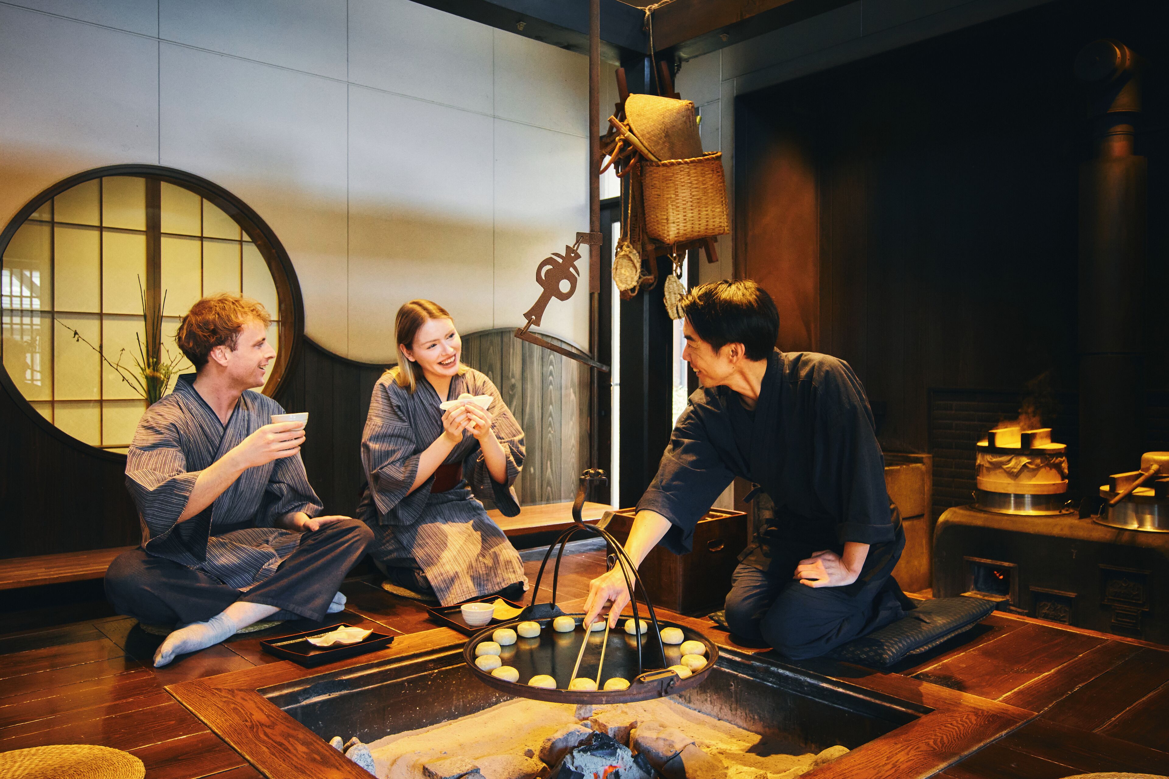Three people sitting on the floor around a traditional Japanese grill, with a wooden floor and a large round mirror in the background.