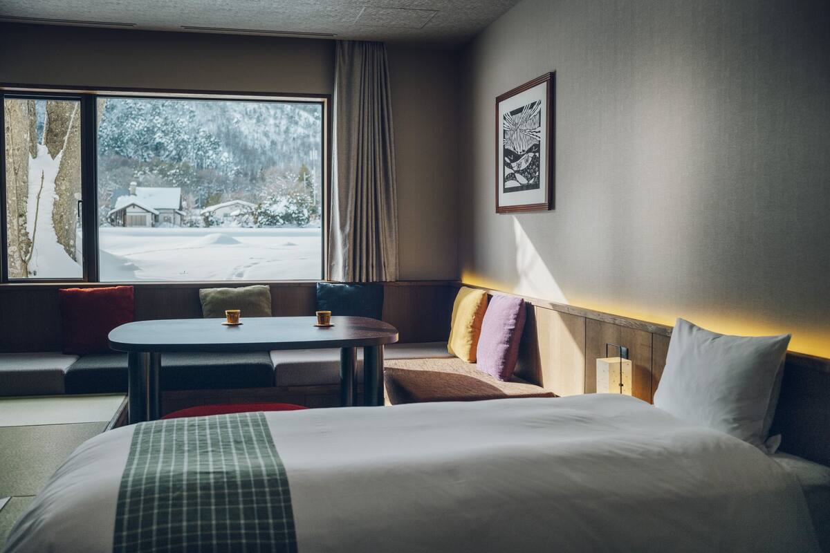 A hotel room with a large bed, a small table, a sofa, and a window with a view of a snowy landscape.