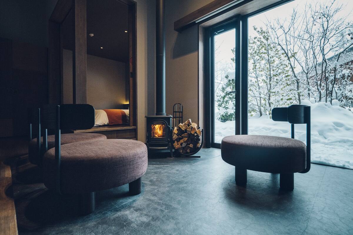 A cozy room with a fireplace, two cushioned chairs, a bed, and a large window overlooking a snowy landscape.