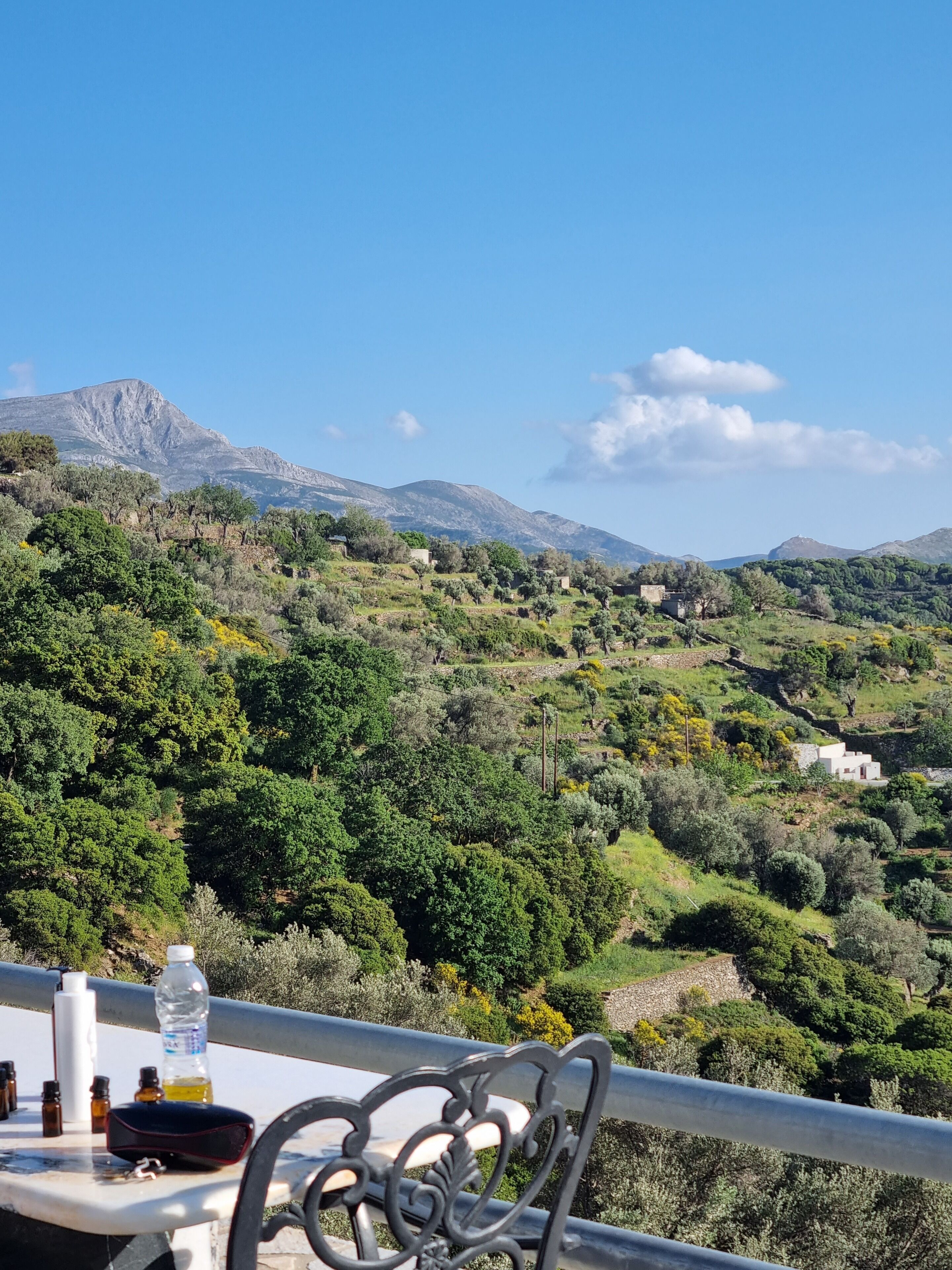 A table with a chair, a bottle of water, and some small bottles on a balcony overlooking a mountainous landscape with scattered buildings.