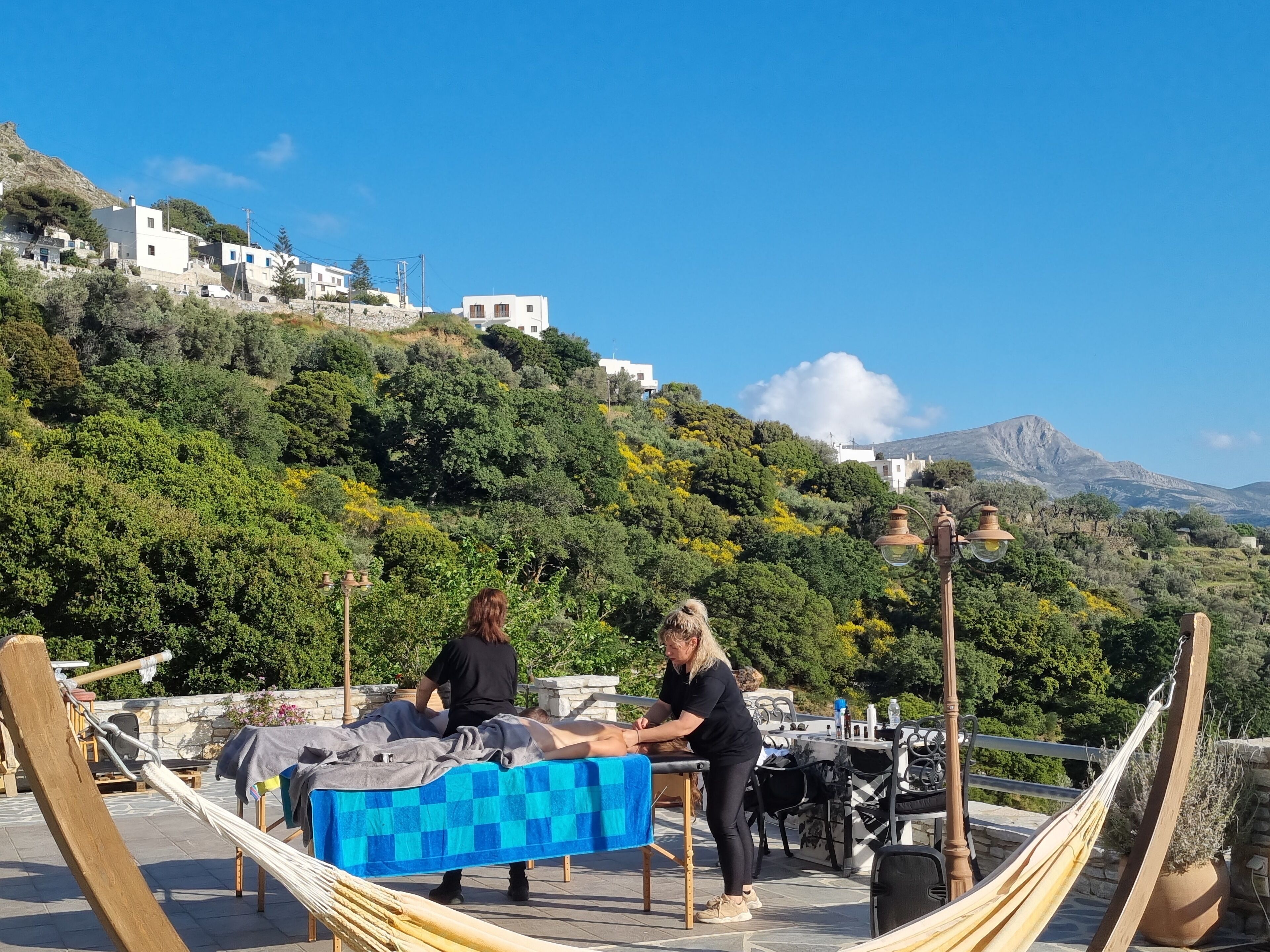 A rooftop terrace with a checkered table, hammocks, and a scenic view of a hillside with buildings.