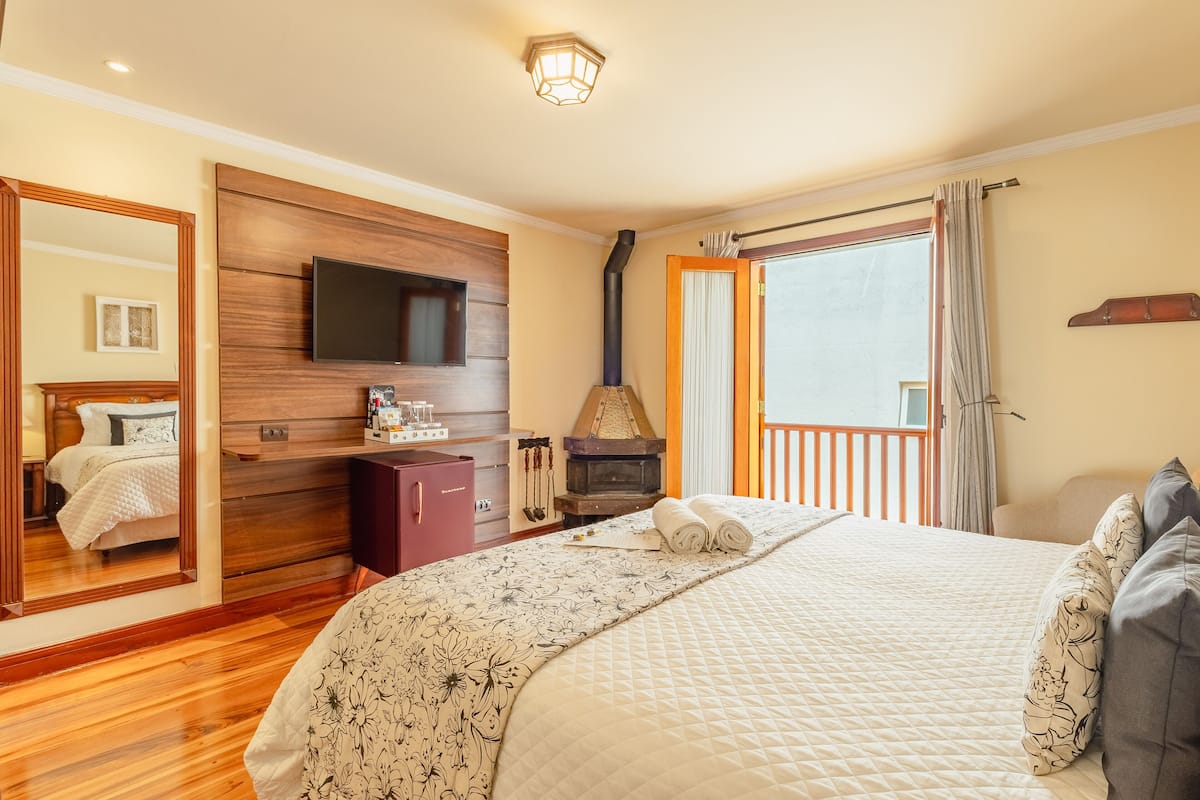 A bedroom with a large bed, a television mounted on a wooden panel, a fireplace, and a balcony with curtains.