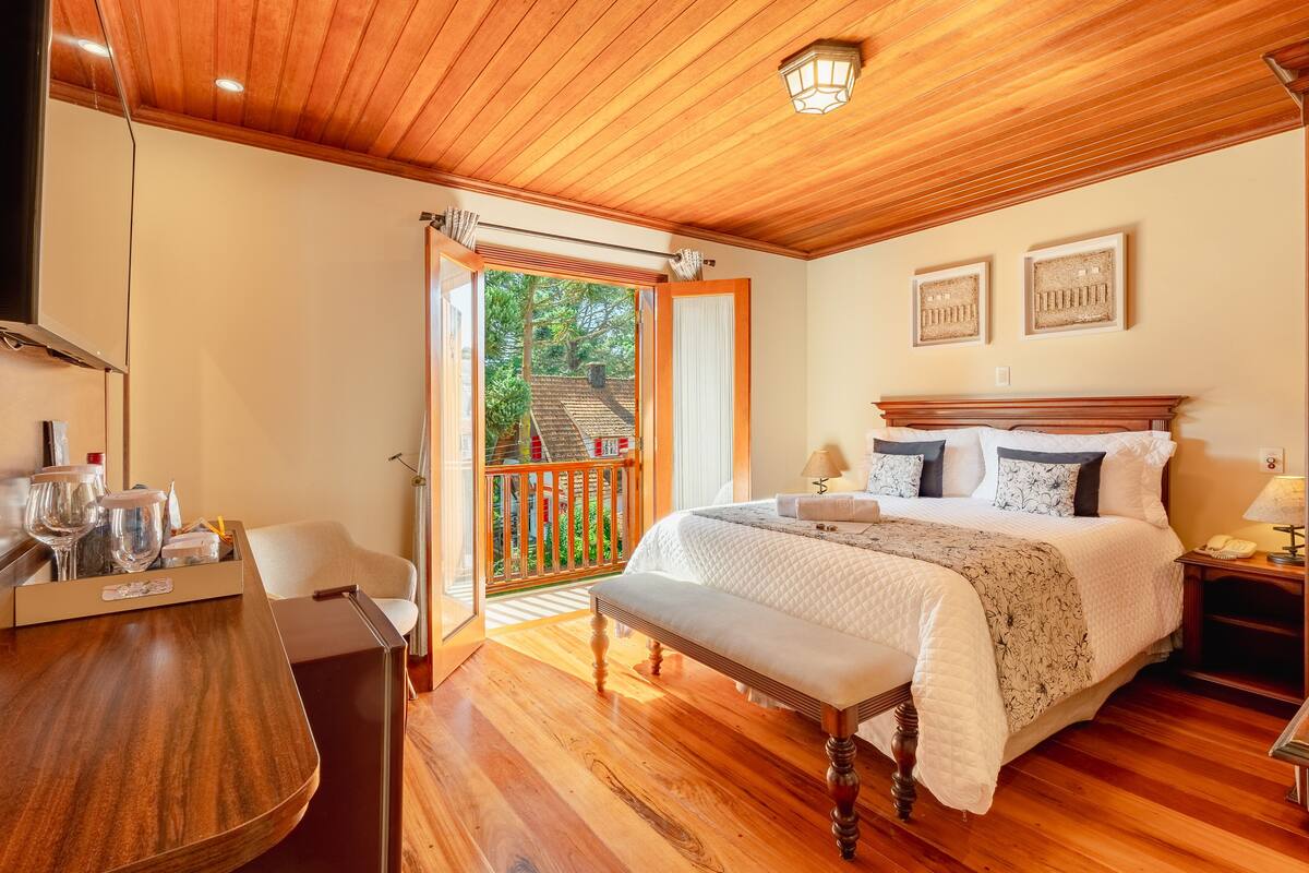 A bedroom with a wooden ceiling, a bed with a white quilt, a bench, a bedside table with a lamp, and a view of a balcony with a railing.