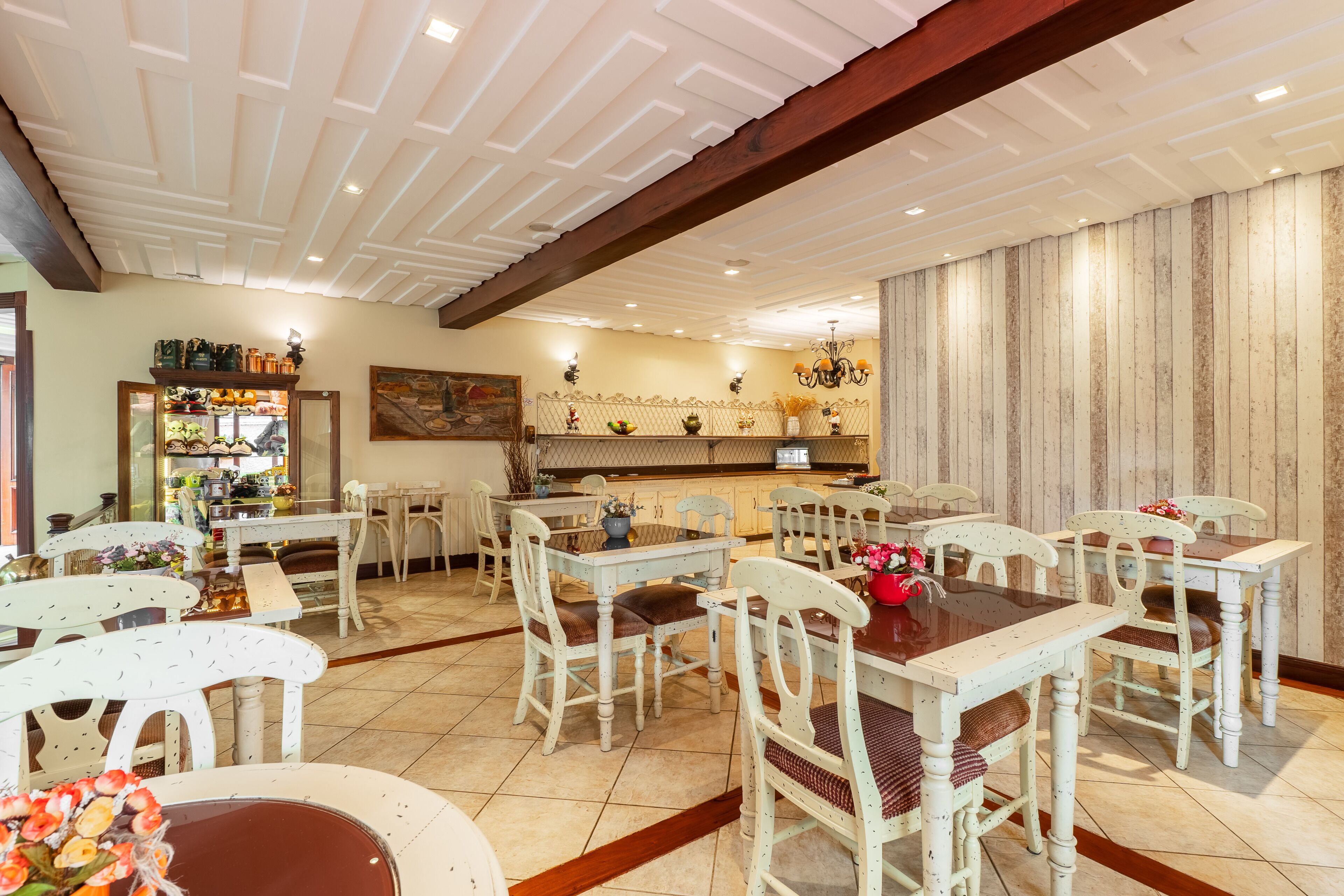 Un espace repas avec des tables et des chaises en bois peintes en blanc, une cuisine à l’arrière-plan et un plafond décoratif.