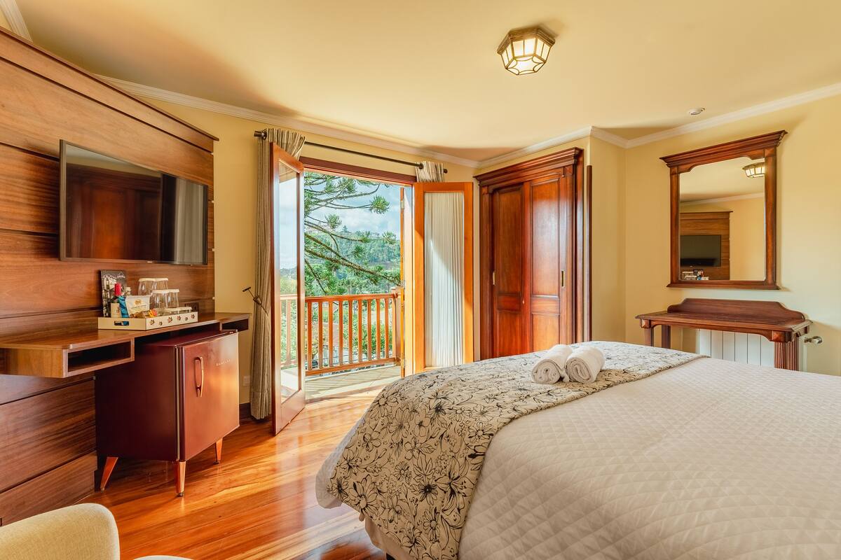 A hotel room with a wooden desk, a bed with a floral bedspread, a large mirror, and a balcony with a view of trees.