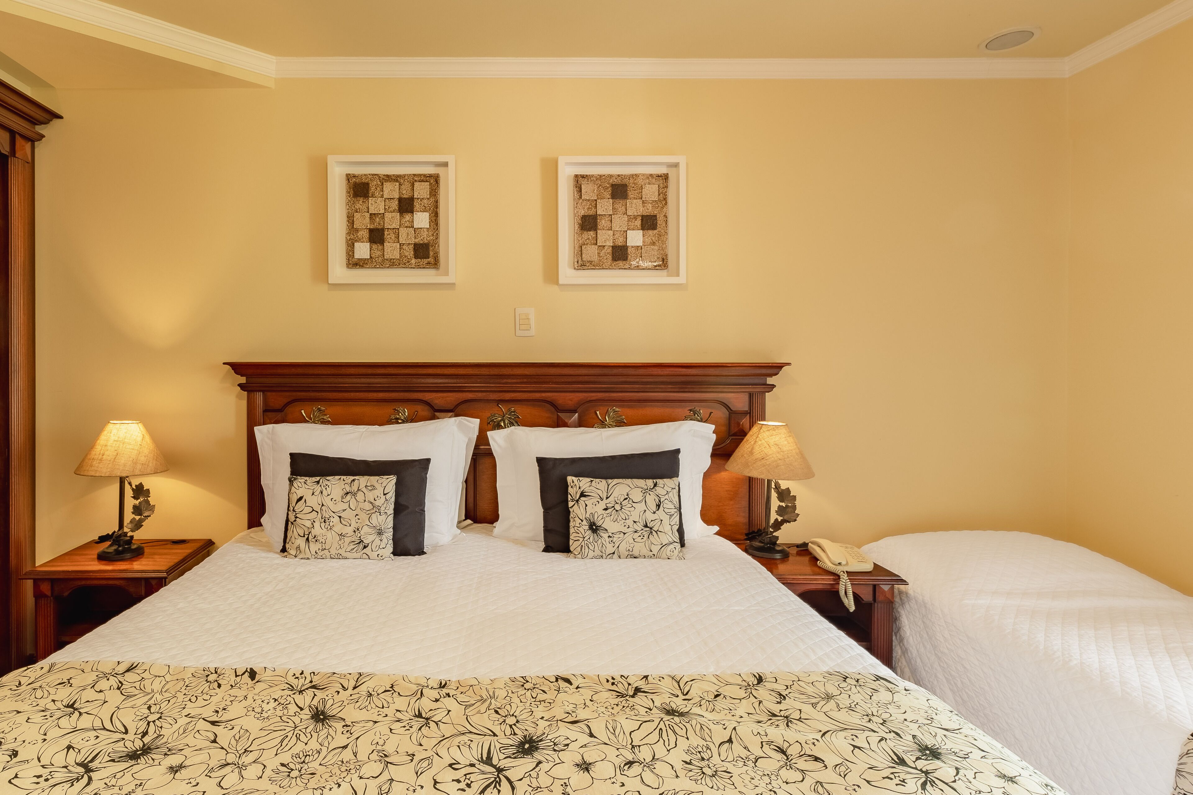 A hotel room with a large bed, two bedside tables, a wooden headboard, two framed artworks on the wall, and a bedside lamp.