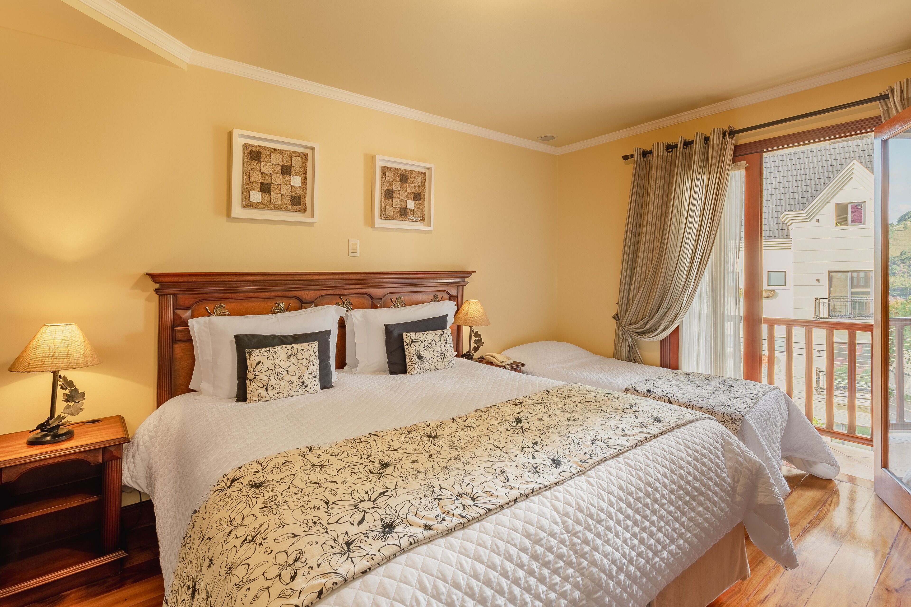 A double bed with a floral bedspread, a wooden nightstand with a lamp, and two framed artworks on the wall.
