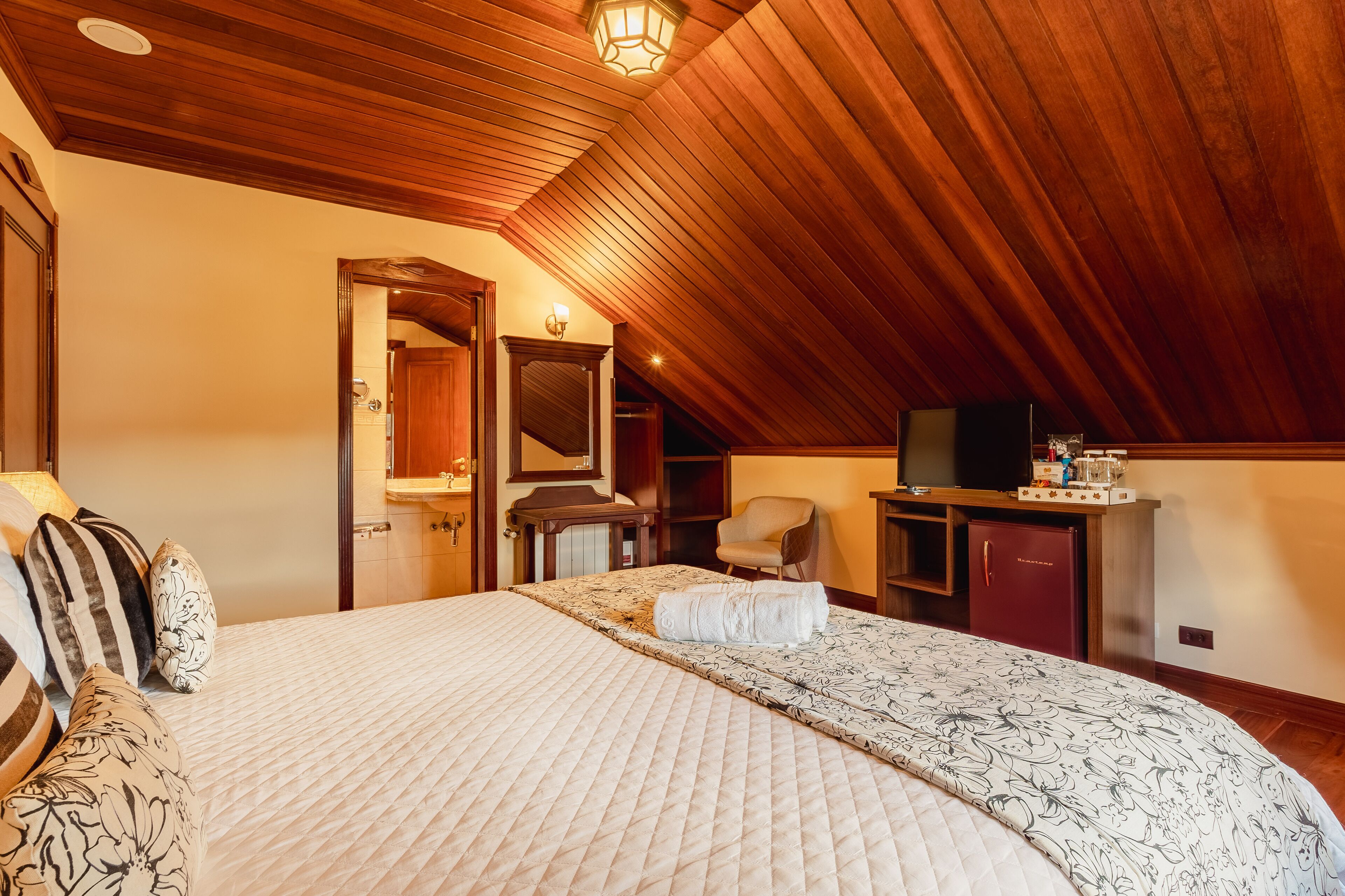 A bedroom with a large bed, a wooden ceiling, a mirror, a chair, a desk, and a television.
