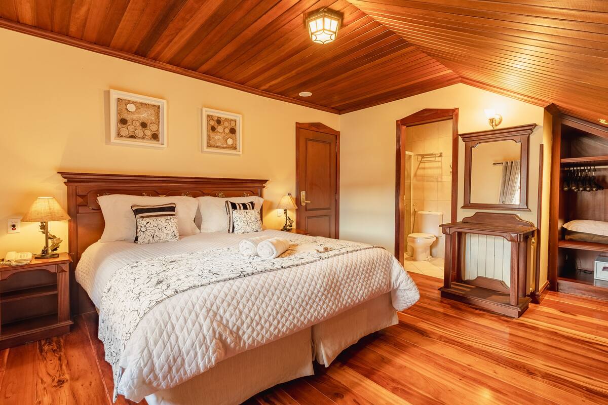 A neatly arranged bedroom with a wooden ceiling, a large bed with a quilted bedspread, two bedside lamps, and a wooden nightstand.