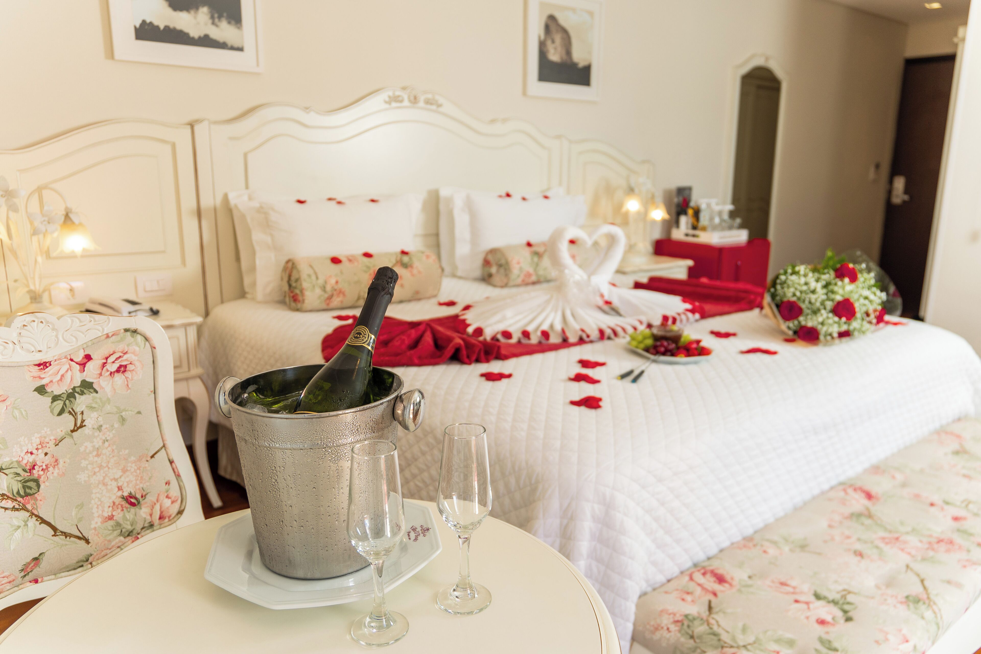 Une chambre d’hôtel avec un lit décoré de pétales de rose, un bouquet de fleurs et un décor en forme de cœur. Une bouteille de champagne se trouve dans un seau sur le lit, et deux verres sont posés sur une table.