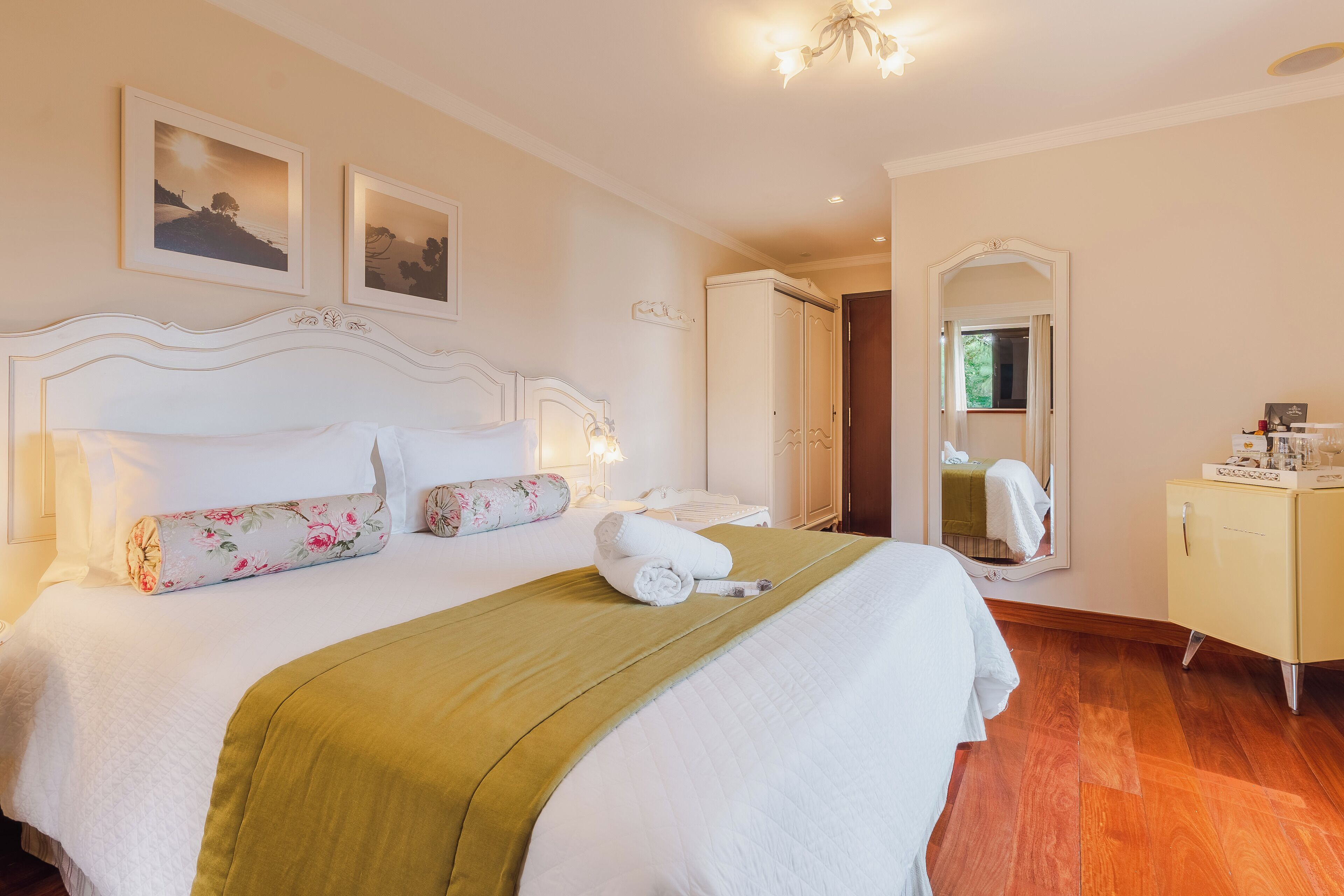A neatly made bed with a floral pillow, a green throw blanket, and a rolled towel. There is a white wardrobe with a mirror and a small table beside the bed.