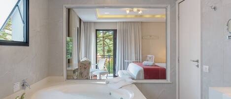 Un baño moderno con una bañera grande, un espejo que refleja un dormitorio con cama y cortinas, y una ventana con vistas a la vegetación.