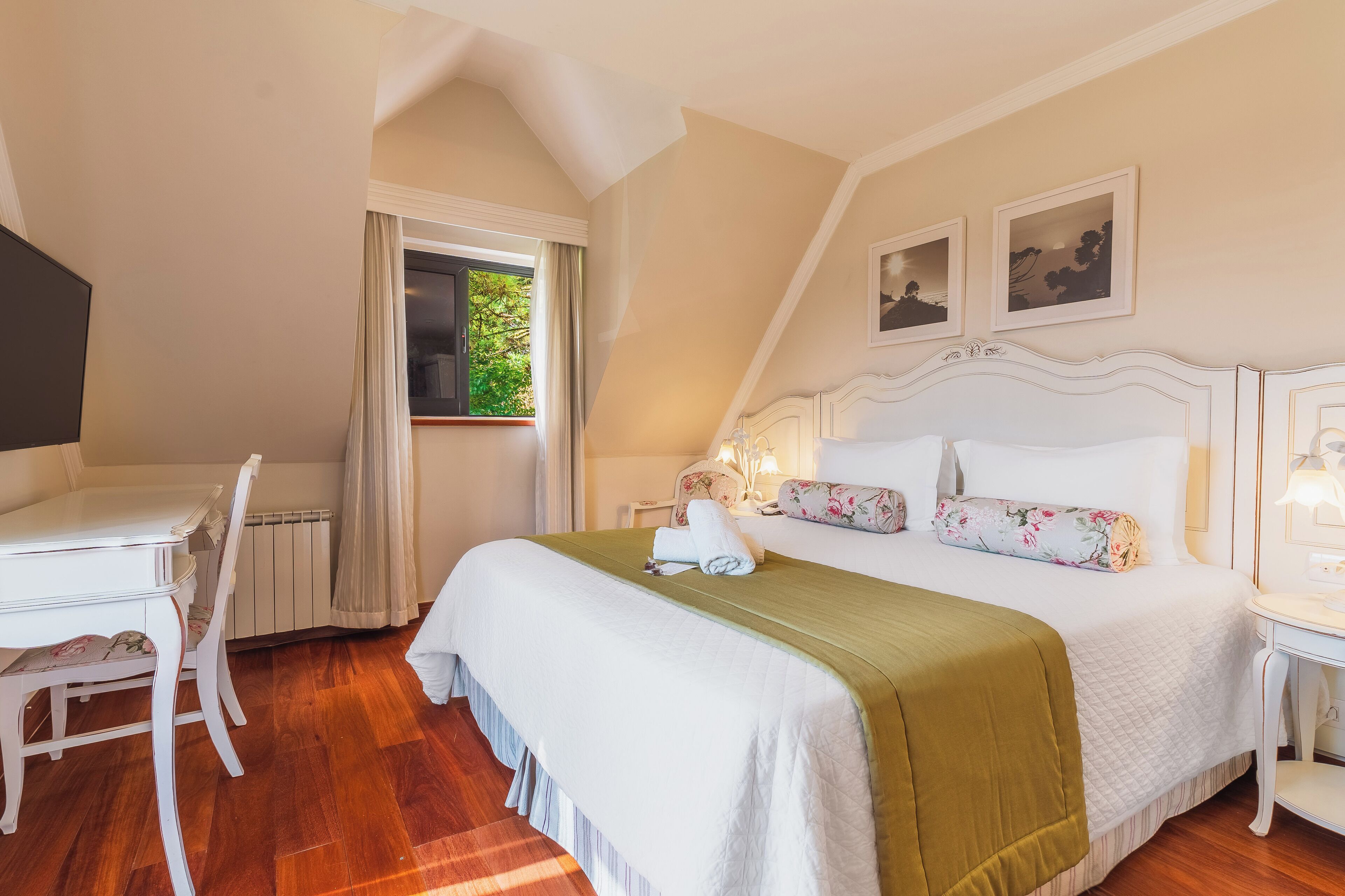A bedroom with a bed, a desk, and a chair. There is a window with curtains, a radiator, and a picture on the wall.