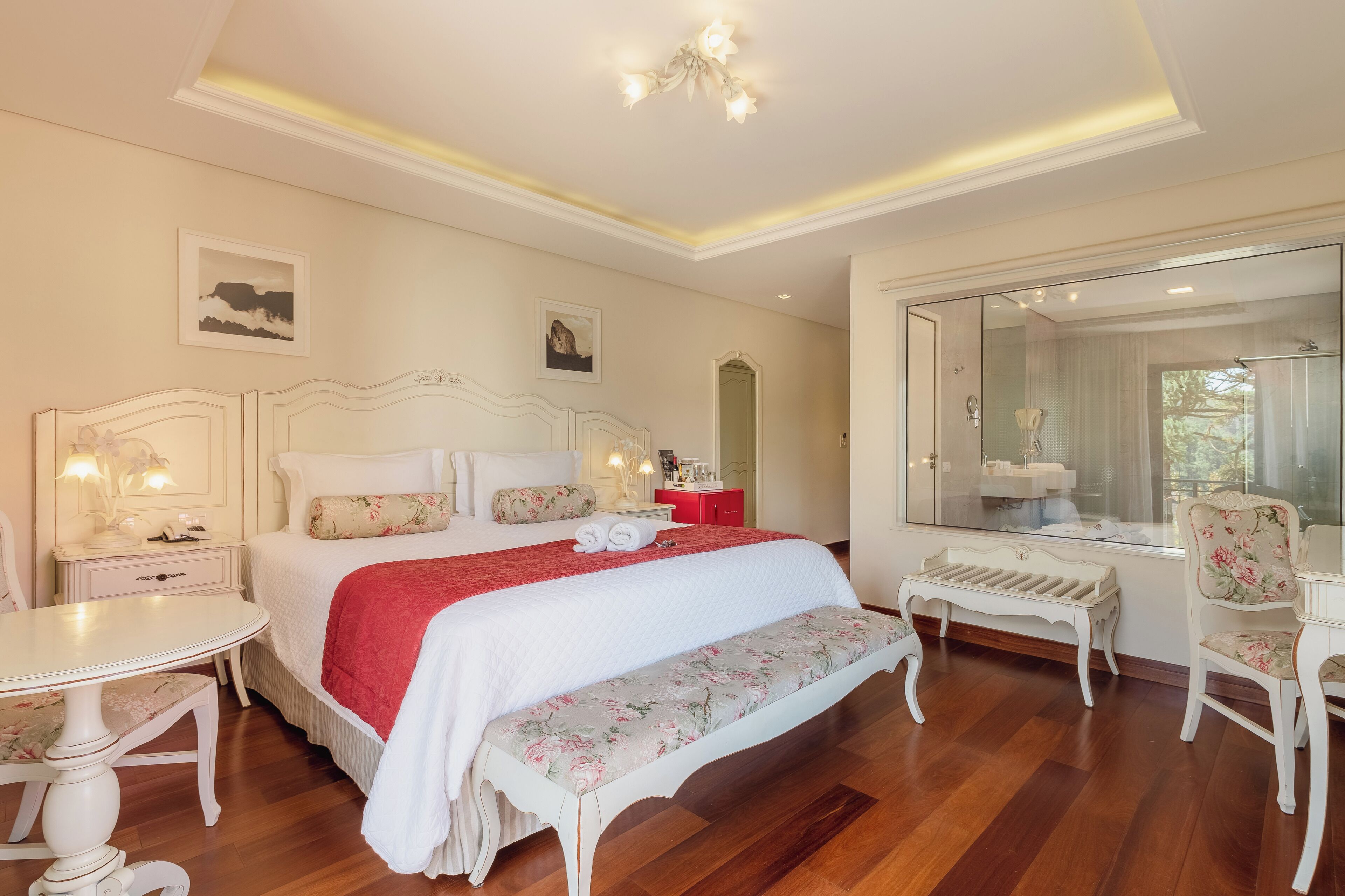 Quarto com uma cama grande, uma mesa de cabeceira, um banco, uma cadeira e um espelho amplo.