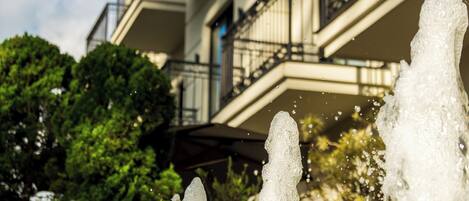 A multi-story building with balconies, a fountain in the foreground, and lush greenery in the background.