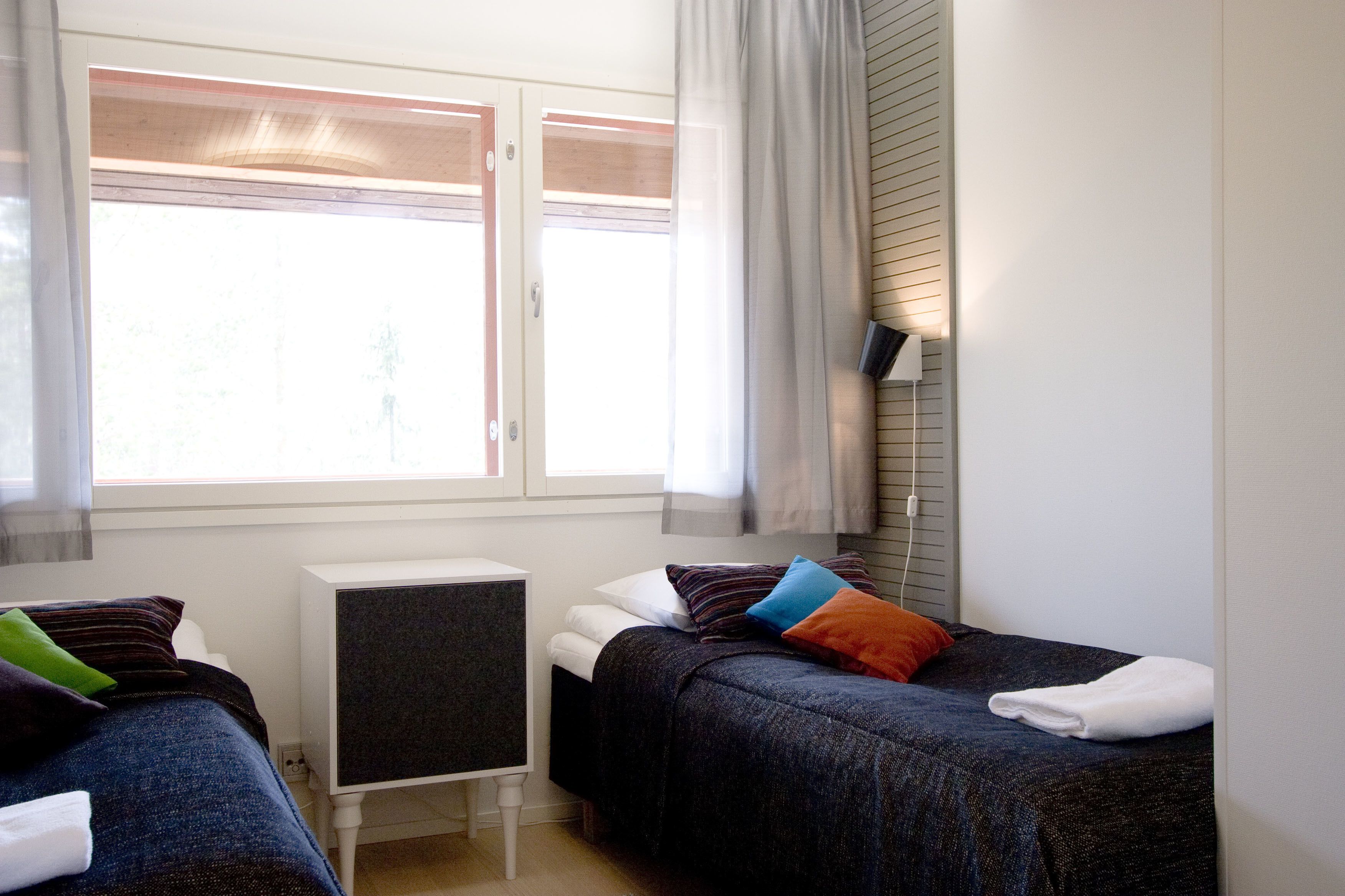 A hotel room with two beds, a small desk, a window with curtains, and a wall-mounted lamp.