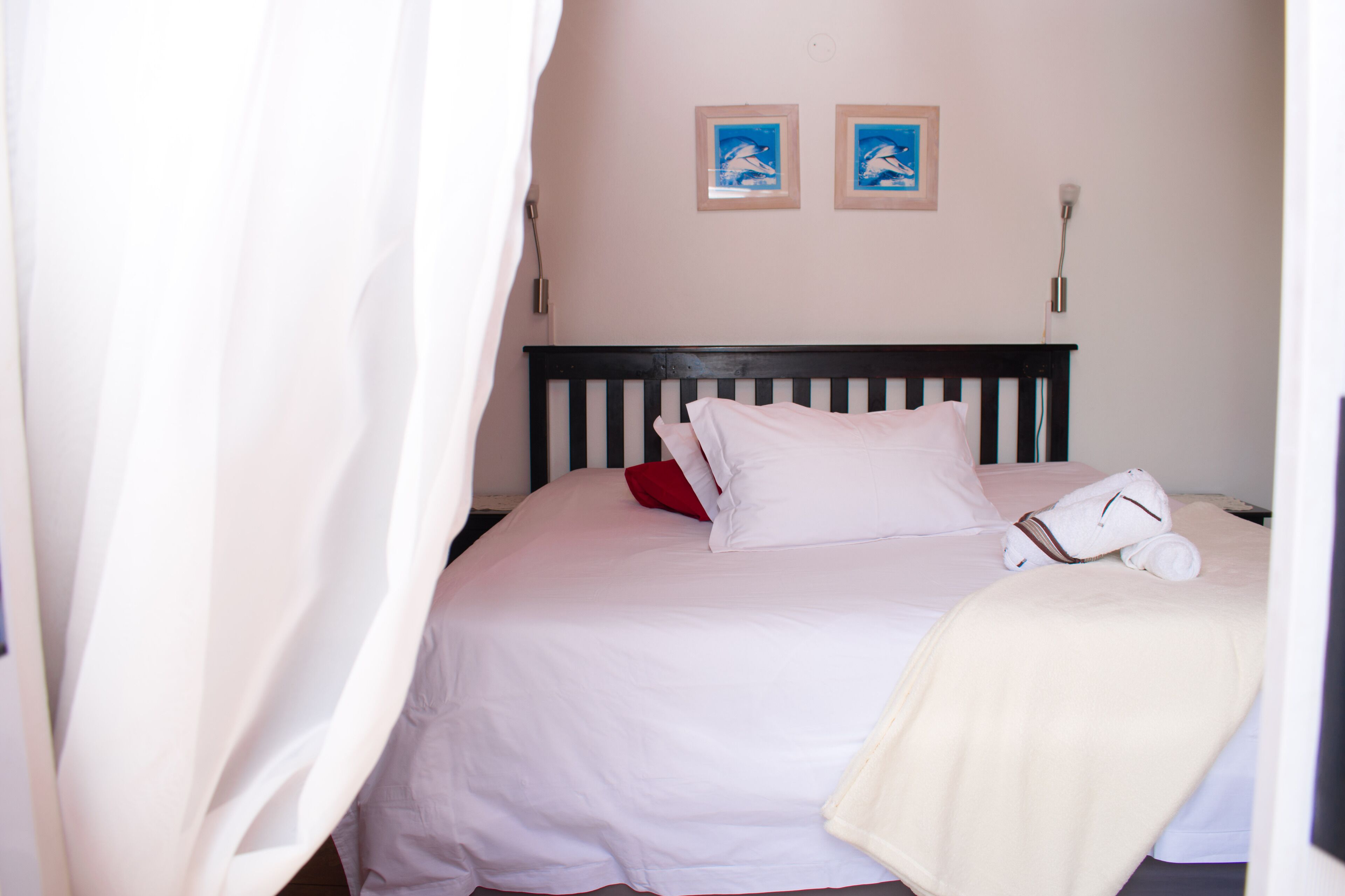 A neatly made bed with white bedding and a red pillow, a white towel, and a beige blanket. There are two framed pictures on the wall above the bed.