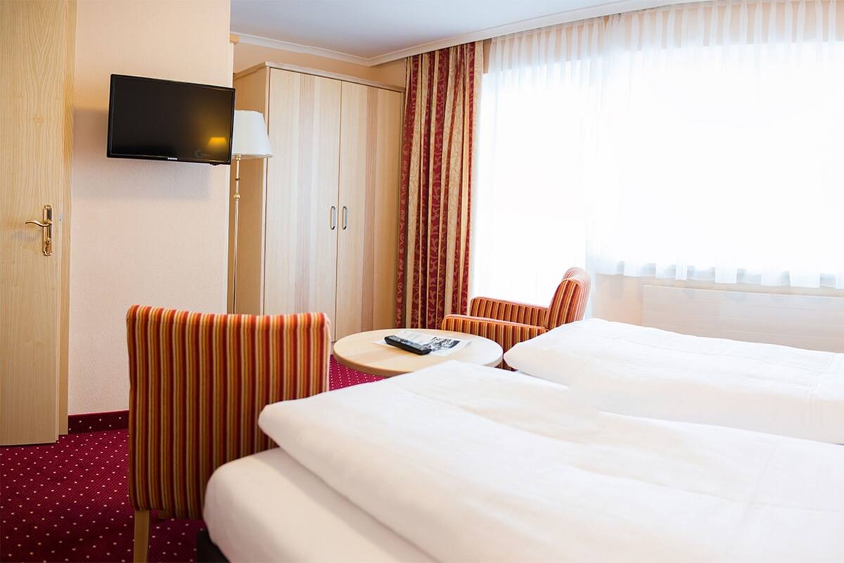 Une chambre d’hôtel avec deux lits, un téléviseur fixé au mur, un bureau avec une chaise et une fenêtre avec des rideaux.