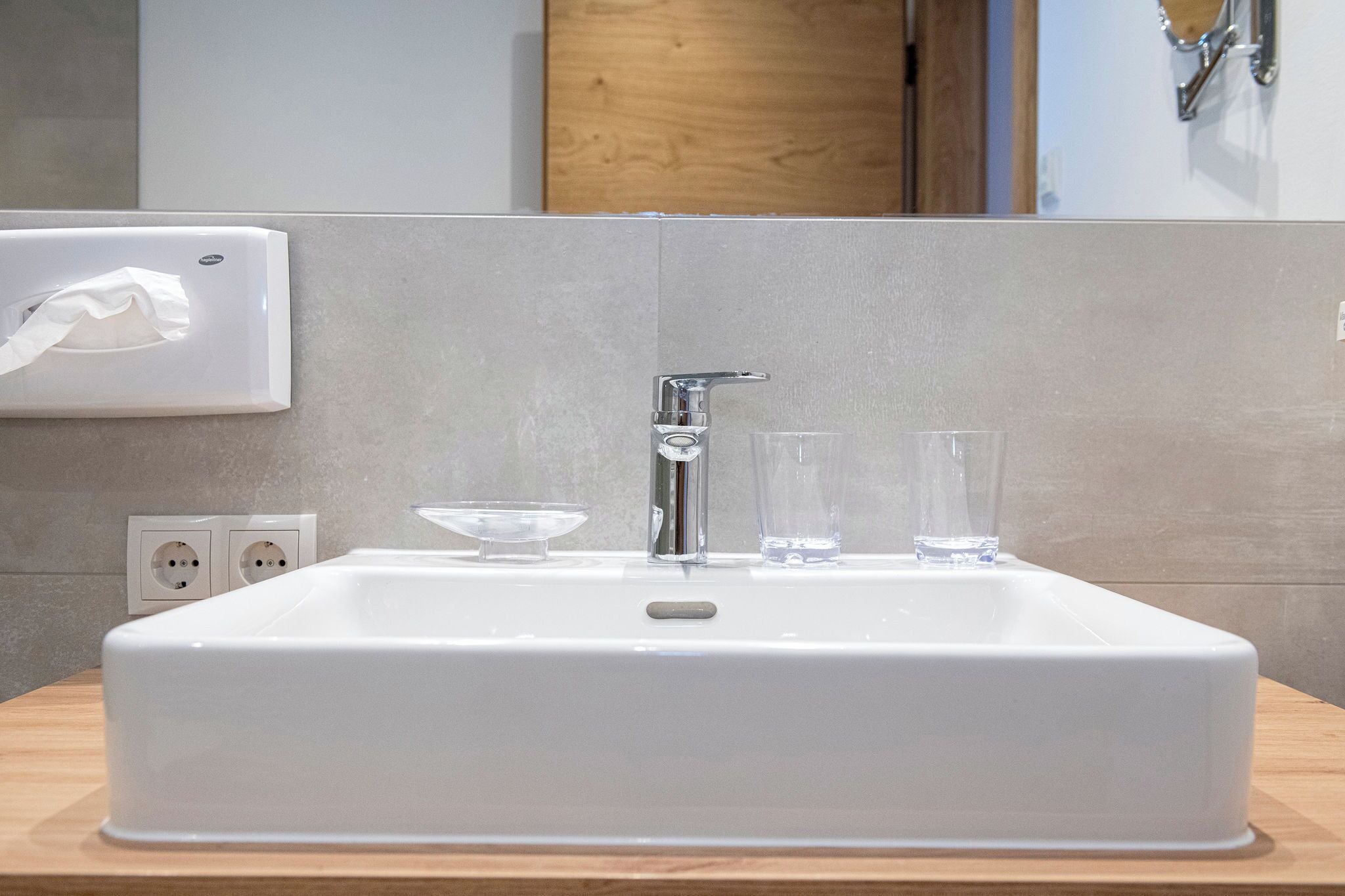 Ein weißes Waschbecken mit einem modernen Wasserhahn, einem Papierspender und zwei klaren Gläsern auf der Arbeitsplatte.