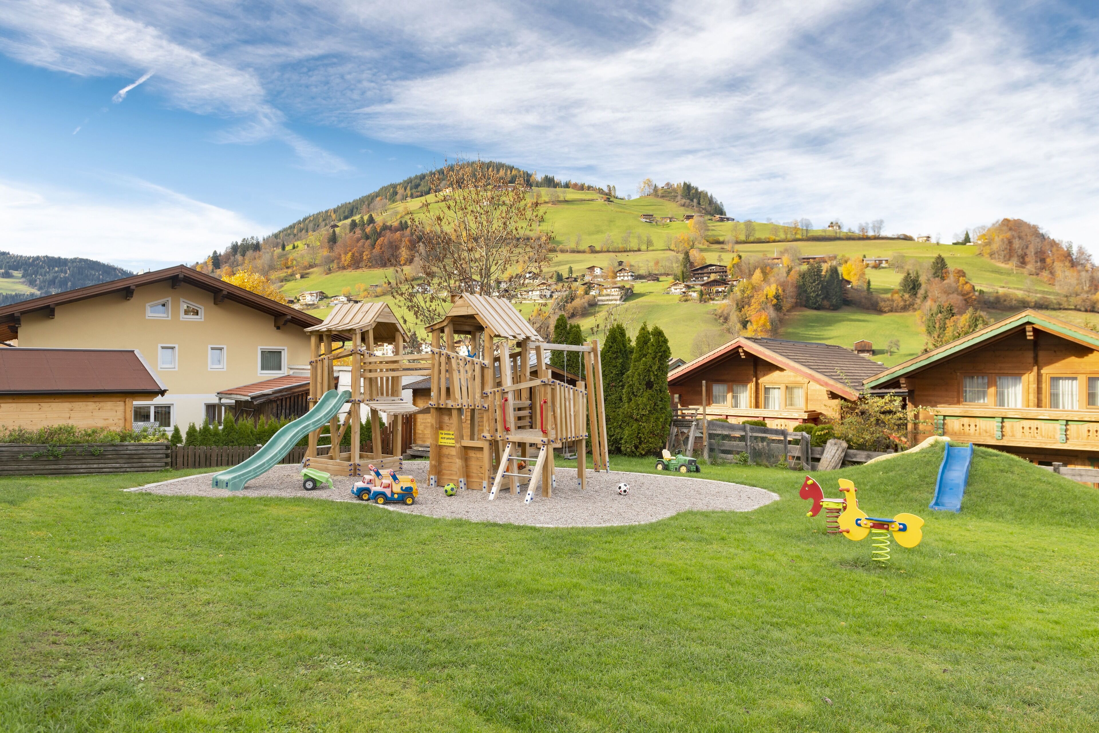 Ein Spielplatz mit Rutschen und Klettergerüsten, umgeben von Häusern und einer hügeligen Landschaft.