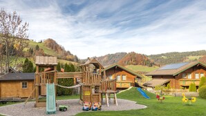 Un parque infantil con tobogán y estructuras para escalar, rodeado de una casita de madera y una casa con tejado inclinado.