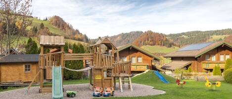 A playground with a slide and climbing structures, surrounded by a wooden playhouse and a house with a sloped roof.