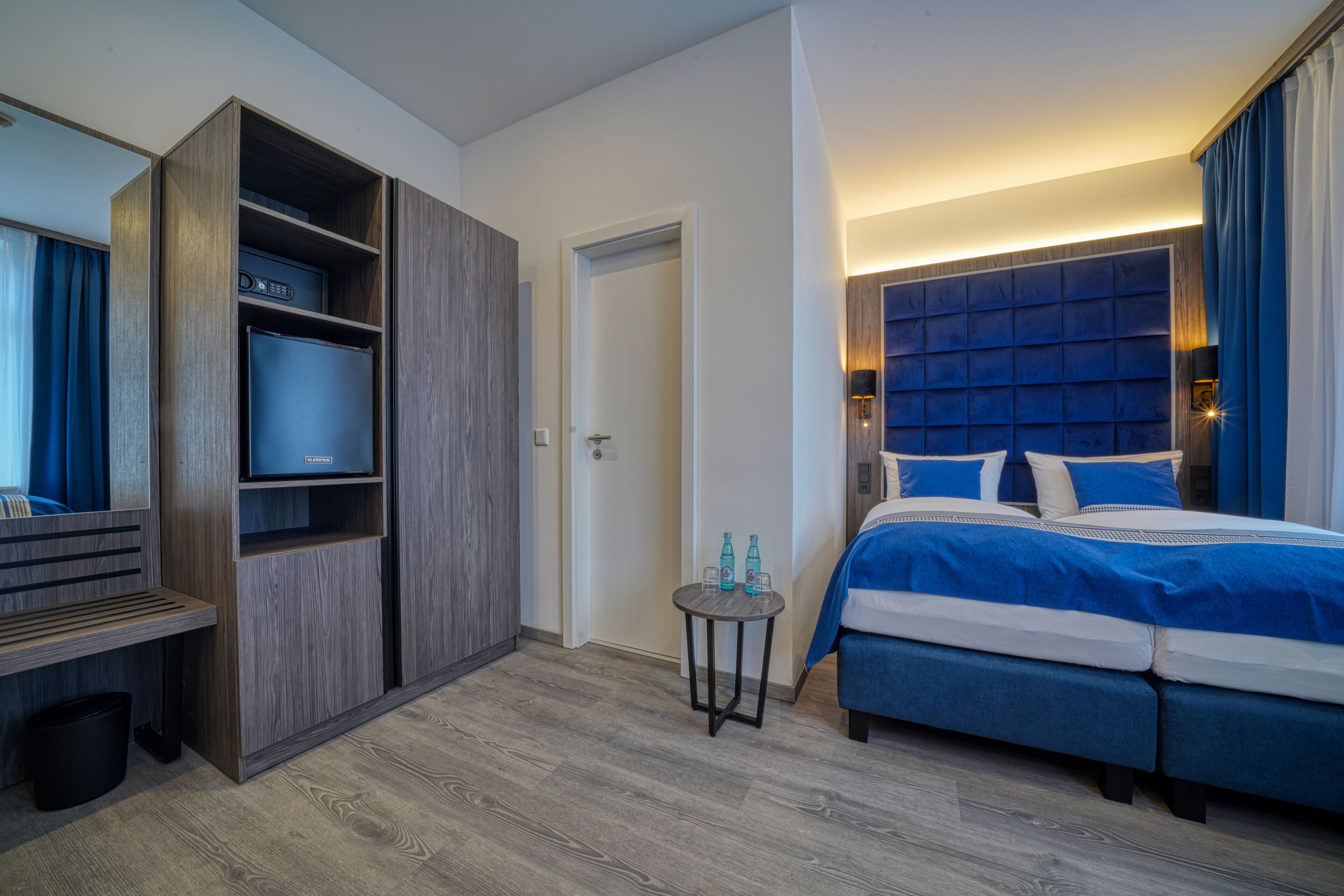 A modern hotel room with a large bed, a built-in wardrobe, a small table with bottles, and a television.