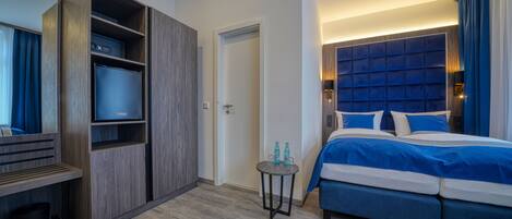 A modern hotel room with a large bed, a built-in wardrobe, a small table with bottles, and a television.