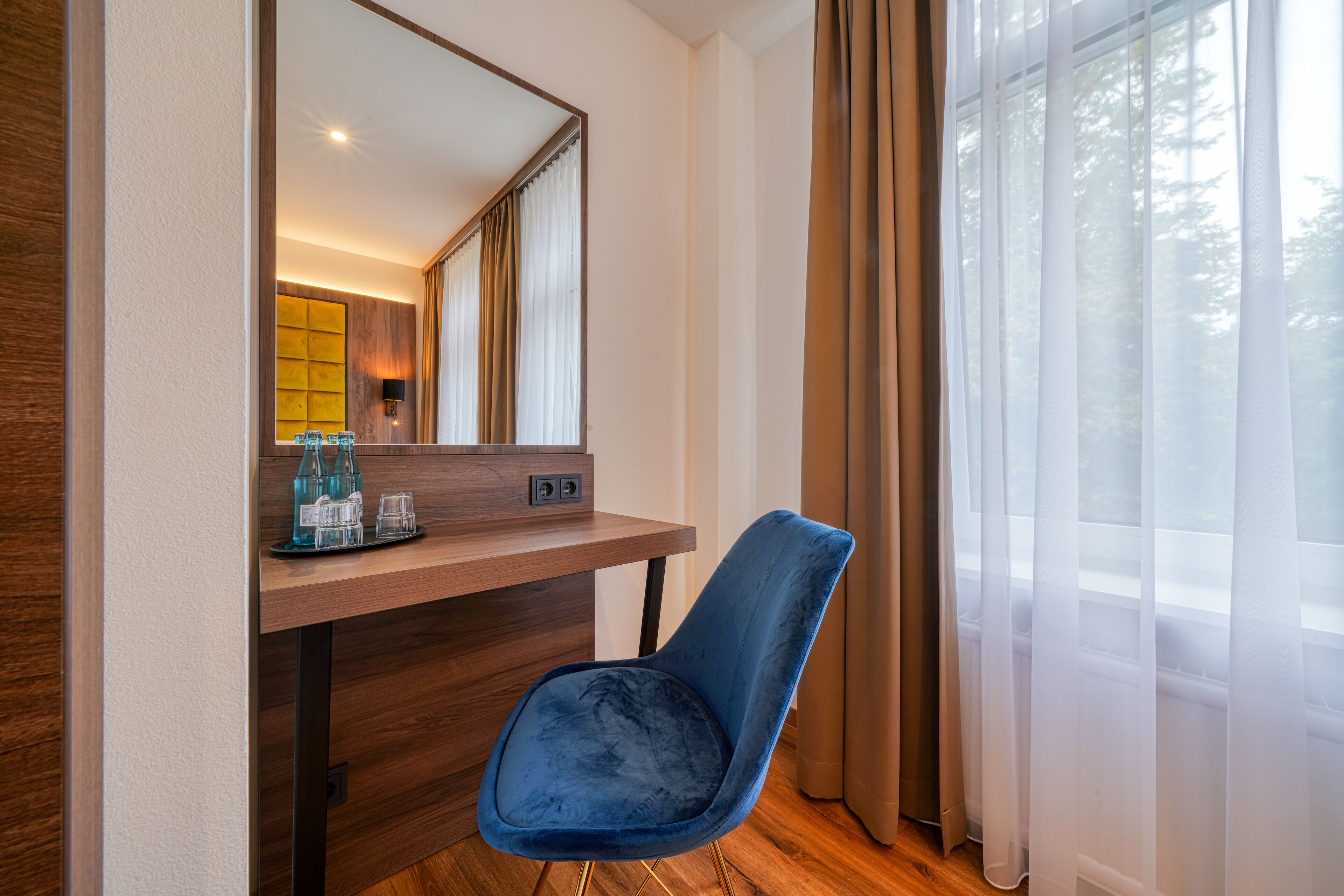 A hotel room with a wooden desk, a blue chair, a mirror, and a window with curtains.