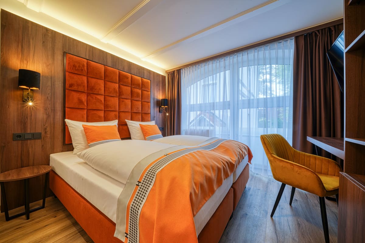 Habitación de hotel con una cama grande, cabecera de madera, dos mesitas de noche, una silla y un ventanal con cortinas.
