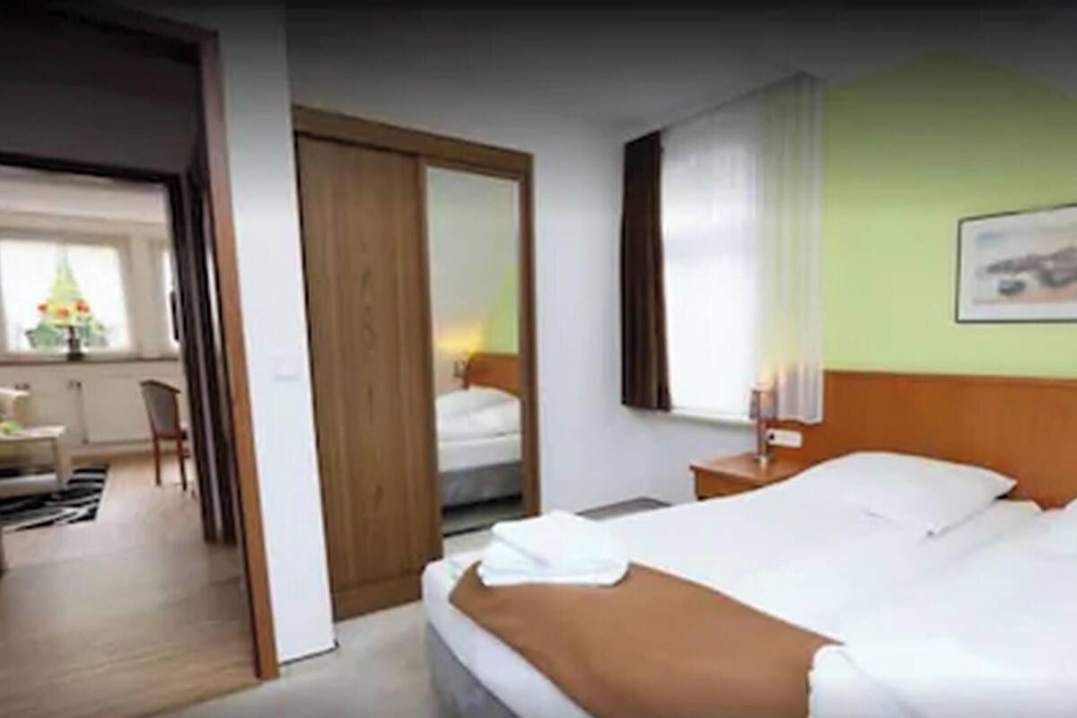 Habitación de hotel con una cama, una mesita de noche, una ventana con cortinas, una silla y una puerta que da a otra habitación.