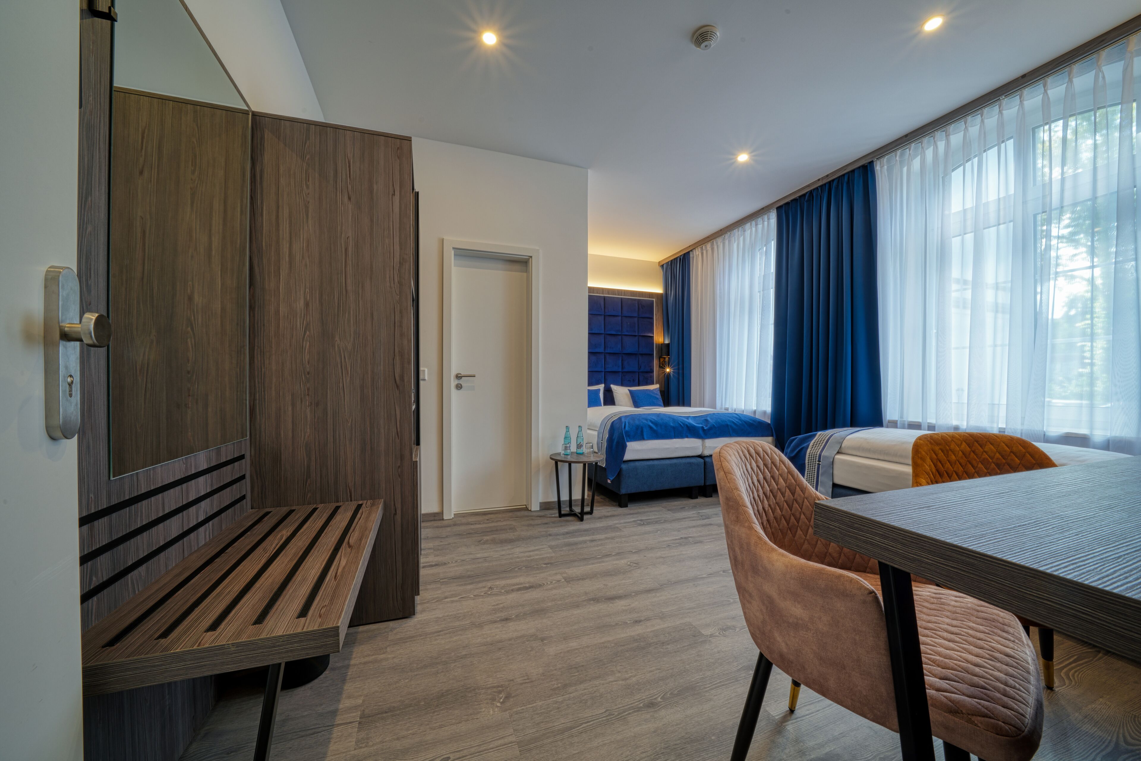 Et moderne hotelværelse med en træbænk, seng med blåt sengetøj, et spisebord med stole og store vinduer med gardiner.