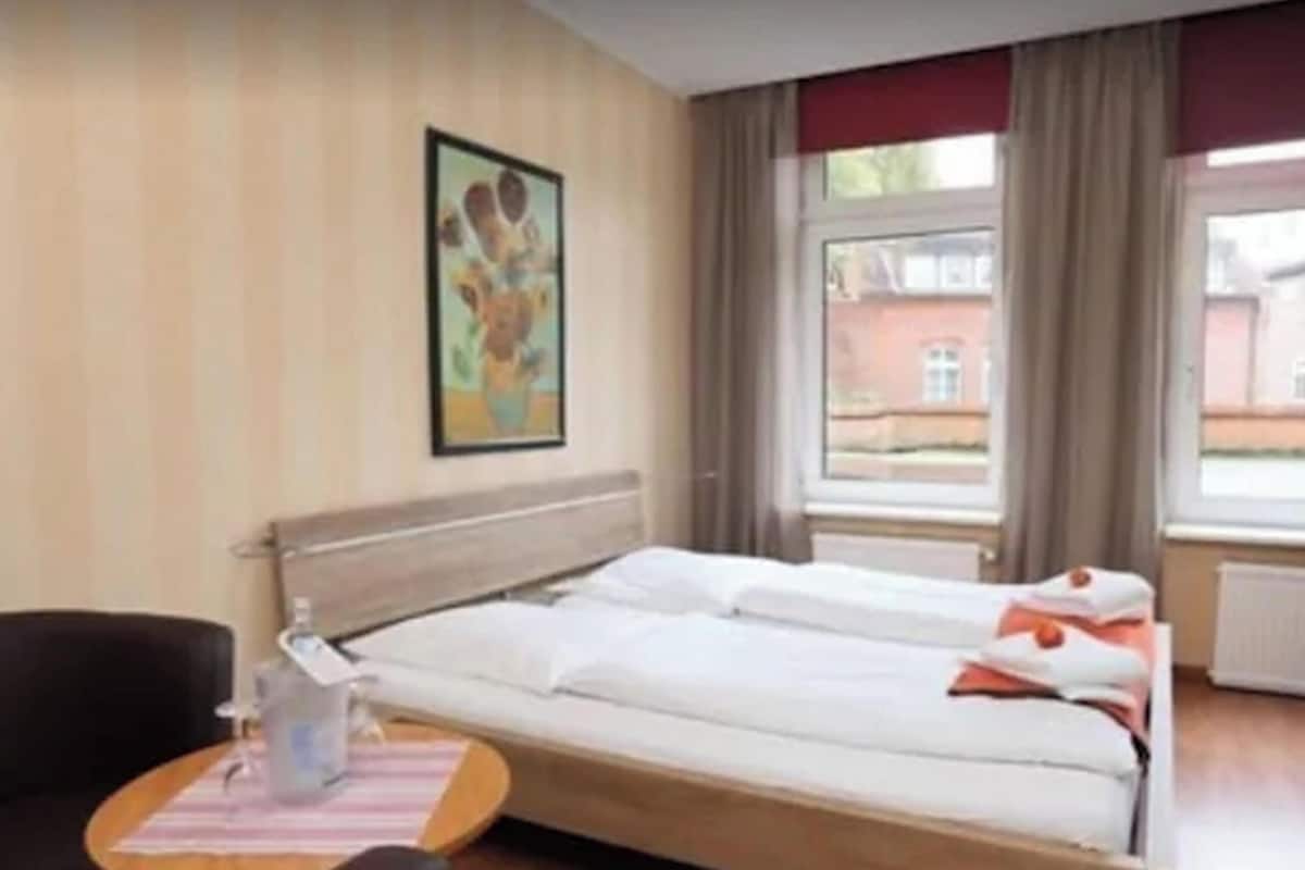 Una habitación de hotel con cama, una silla, una mesita con una botella y una ventana con cortinas.