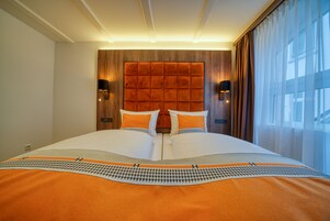 A hotel room with a large bed, wooden headboard, two wall-mounted lamps, and a view of the city through sheer curtains.