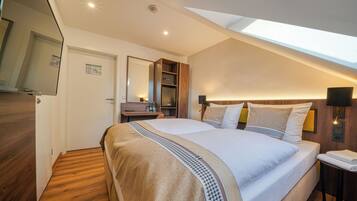 Quarto de hotel moderno com uma cama grande, piso de madeira, claraboia, escrivaninha com cadeira e uma mesinha de cabeceira com abajur.
