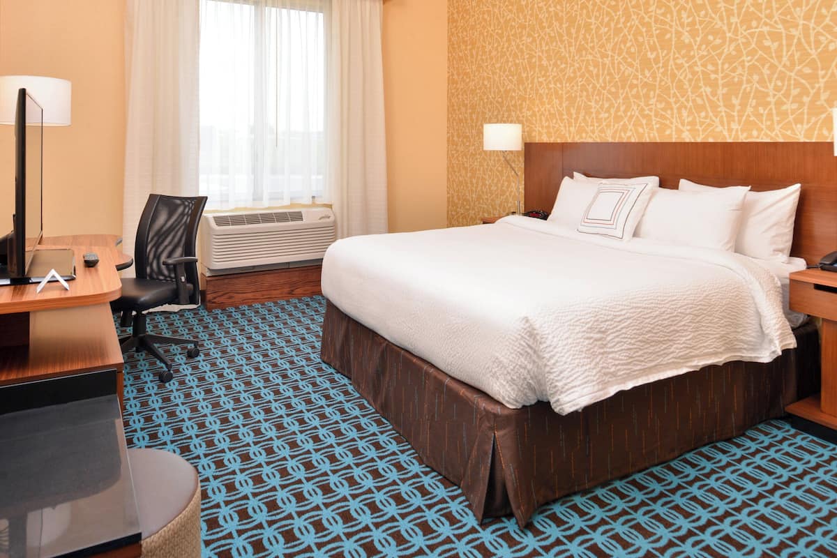 A hotel room with a large bed, a desk with a lamp, a chair, and an air conditioning unit.