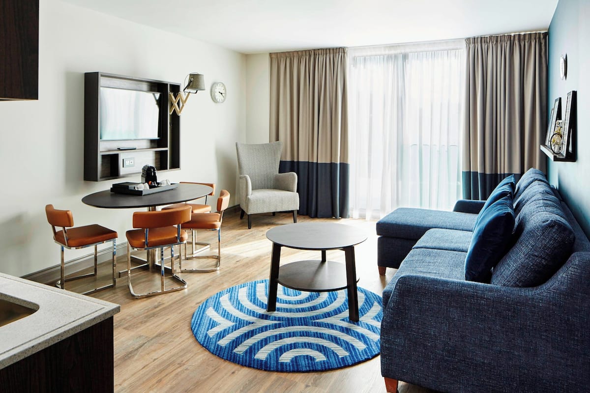 A modern living room with a blue sofa, a round coffee table, a TV mounted on the wall, and a dining area with a table and chairs.