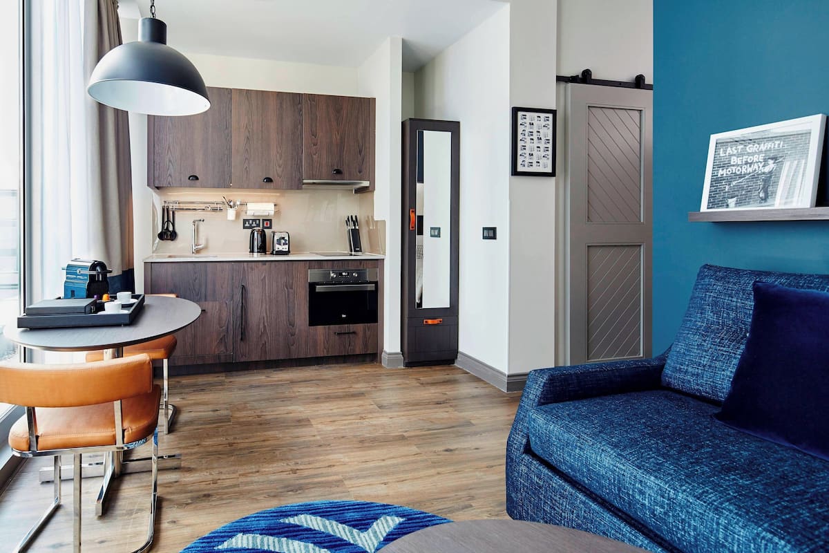 A modern kitchen with wooden cabinets, a dining table with orange chairs, and a blue sofa in a living area with a blue wall and a framed picture.