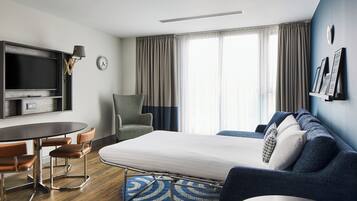 A modern hotel room with a bed, a desk with chairs, a TV, and a large window with curtains.
