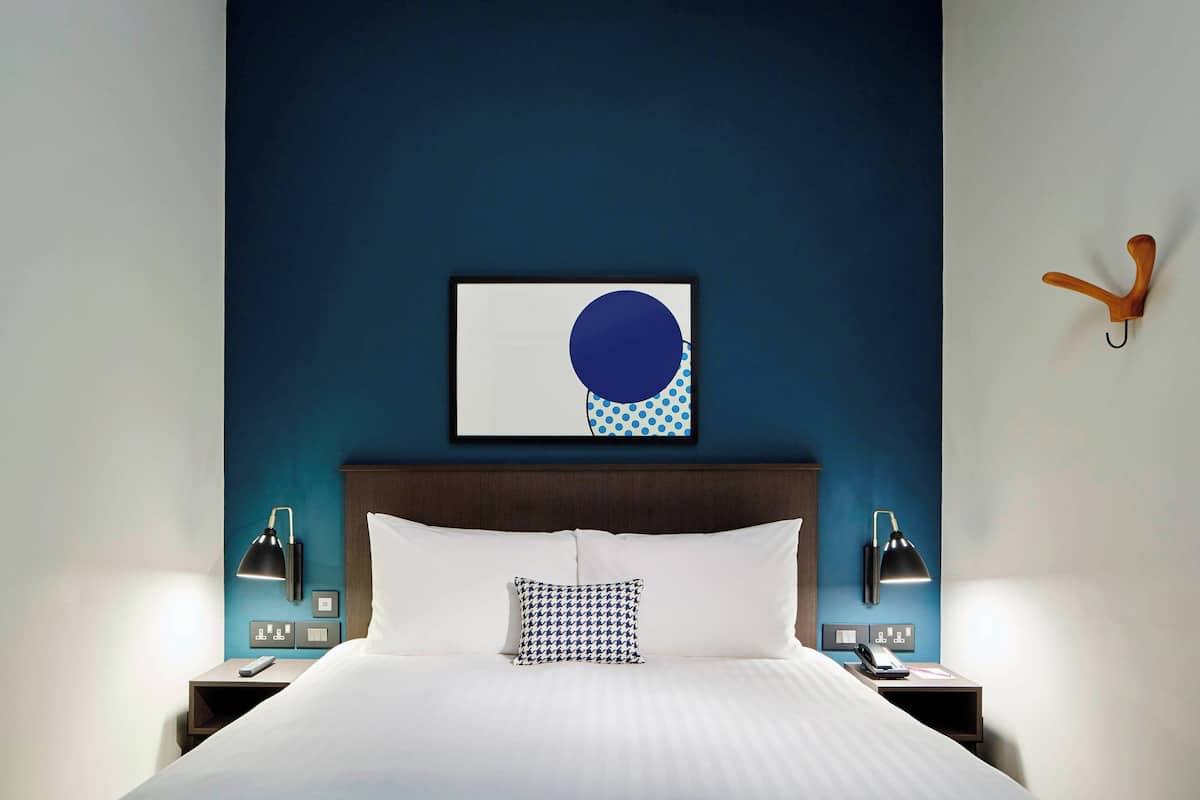 A hotel room with a large bed, two bedside tables, a wall-mounted clock, and a framed artwork above the bed.
