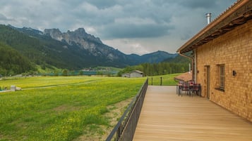 Eine Holzterrasse führt zu einem Gebäude mit Backsteinfassade, mit Blick auf ein grünes Feld und Berge unter bewölktem Himmel.