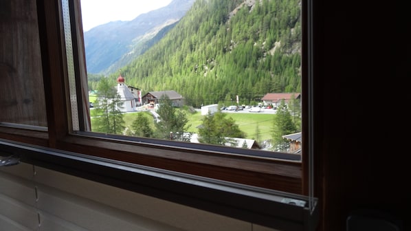 Vista a través de una ventana que muestra un edificio con montañas al fondo, una iglesia y un estacionamiento.