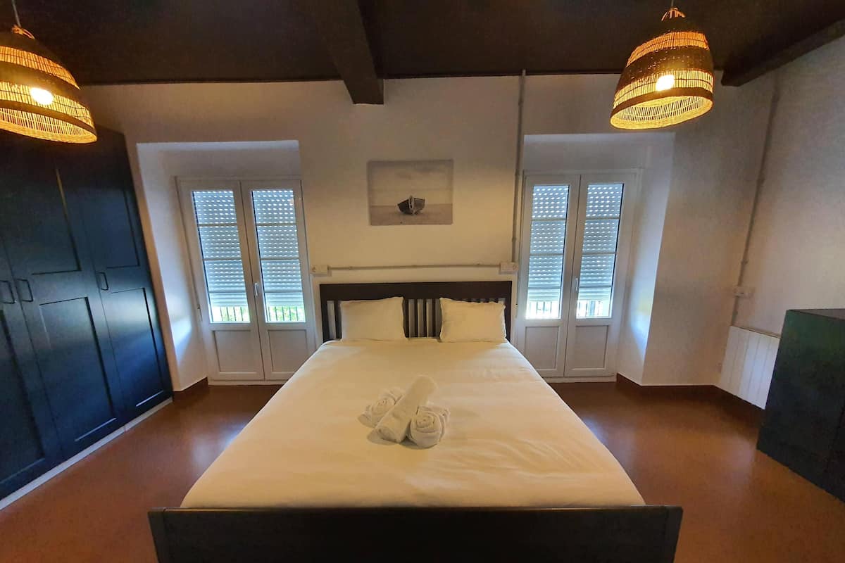 Una cama bien hecha con sábanas y toallas blancas, dos lámparas colgantes, un cabecero oscuro y un pequeño cuadro en la pared.